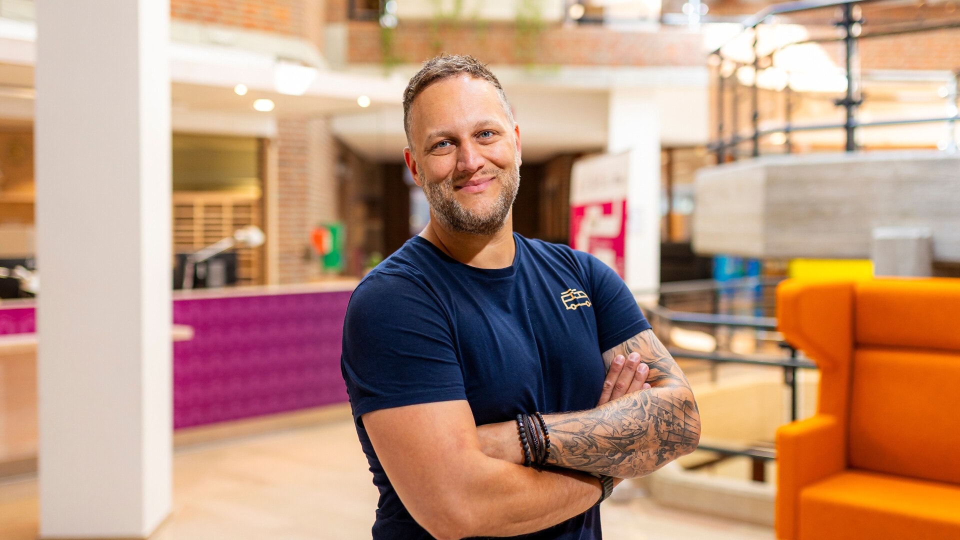 Een lachende man met baard en tatoeages op zijn armen staat met gekruiste armen in een modern kantoorgebouw.