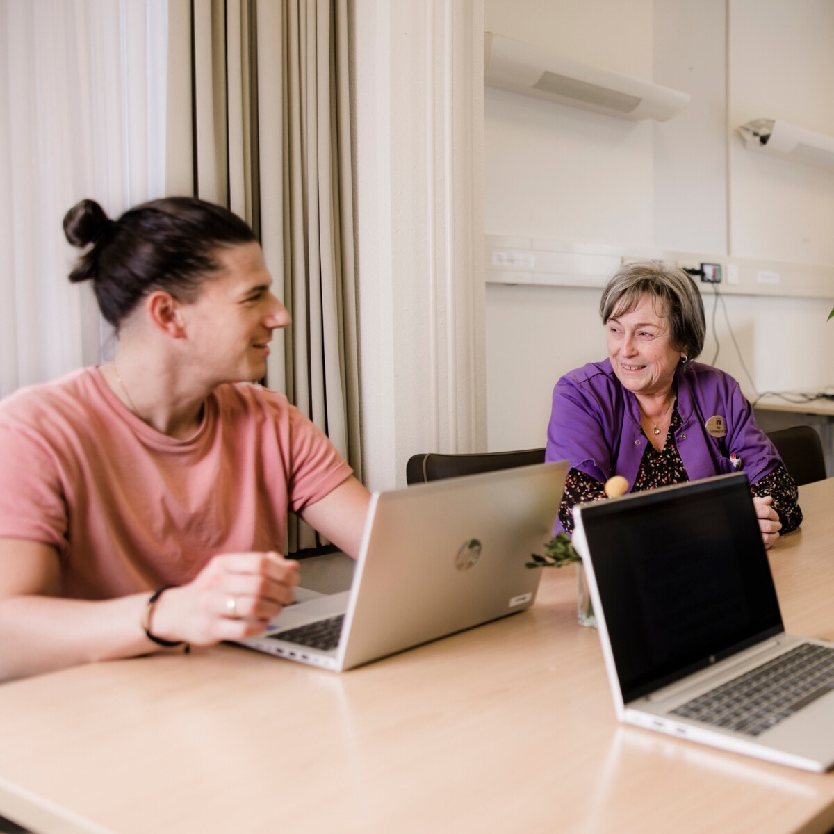 Een man en een oudere vrouw zitten aan een houten tafel in een kantoor. Beiden kijken lachend naar elkaar. De man heeft een laptop voor zich staan.