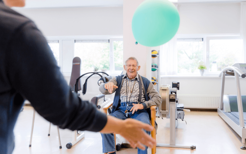 Lachende oudere man die in een stoel zit in een fysioruimte. Hij heeft tegen een lichtblauwe ballon gemept die nu die in de lucht zweeft.