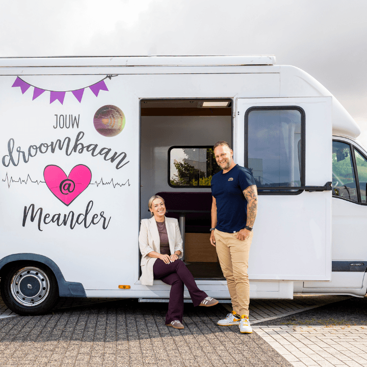 Jonge man en vrouw poseren lachend voor een witte bestelbus met de tekst 'Jouw droombaan @ Meander'.