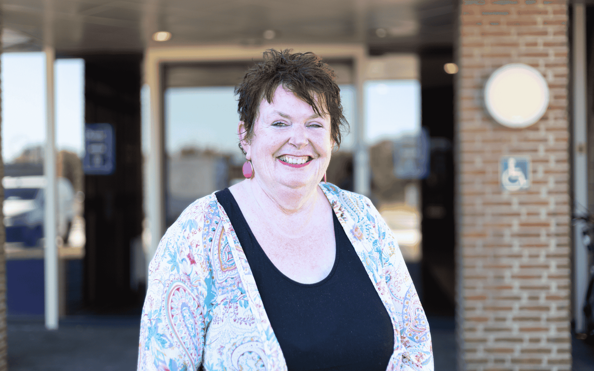 Portret van een lachende vrouw met kort donker haar, een zwarte top en een licht vest met een kleurrijk patroon, staand voor een gebouwingang met glazen deuren en een handicapteken op de muur.