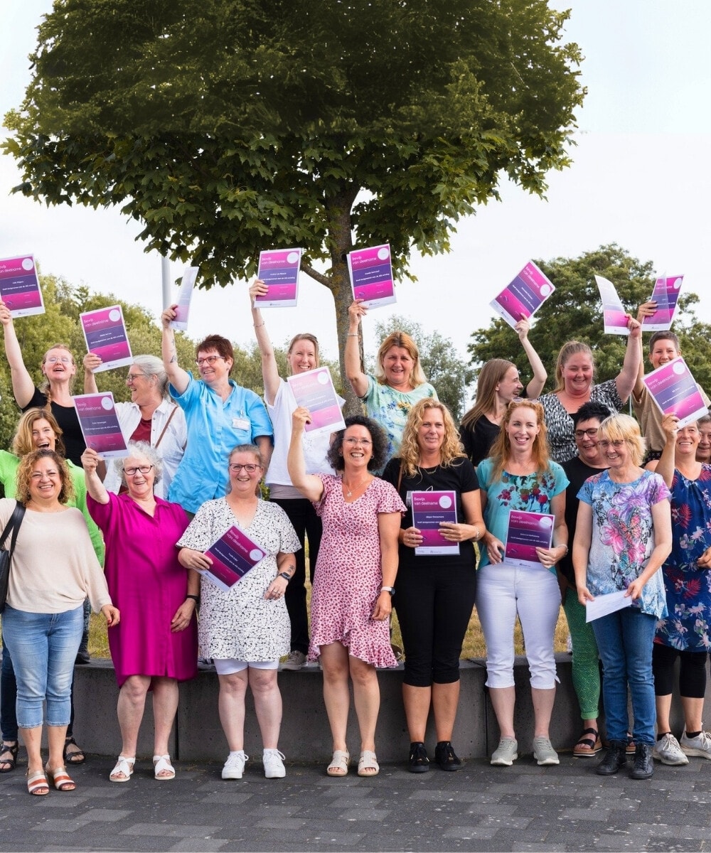 Een grote groep volwassen vrouwen staan buiten met hun armen in de lucht, elk met een paars en roze diploma.