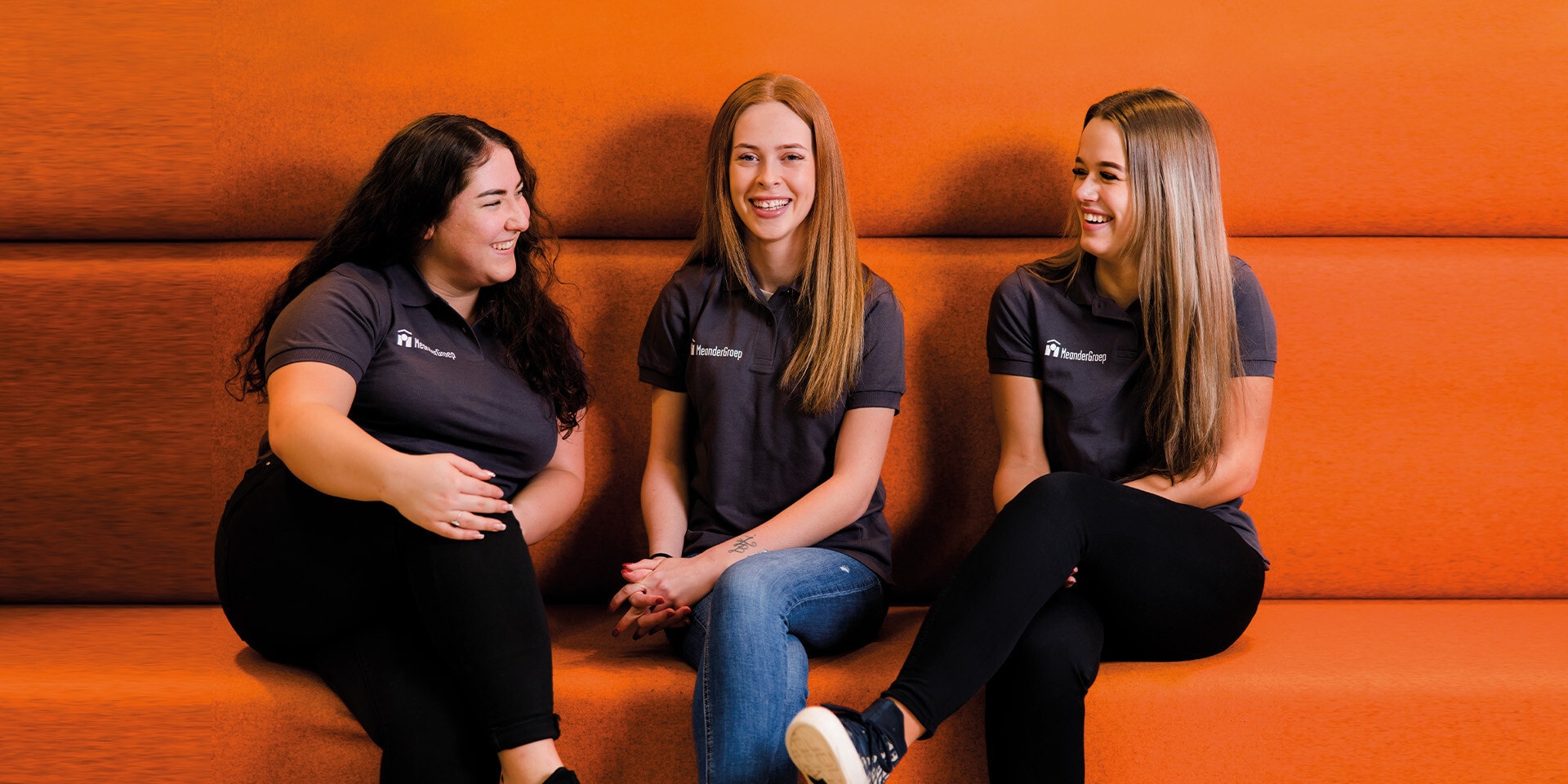 Drie jonge vrouwen zitten op een oranje bank. Ze dragen allemaal een polo van MeanderGroep.