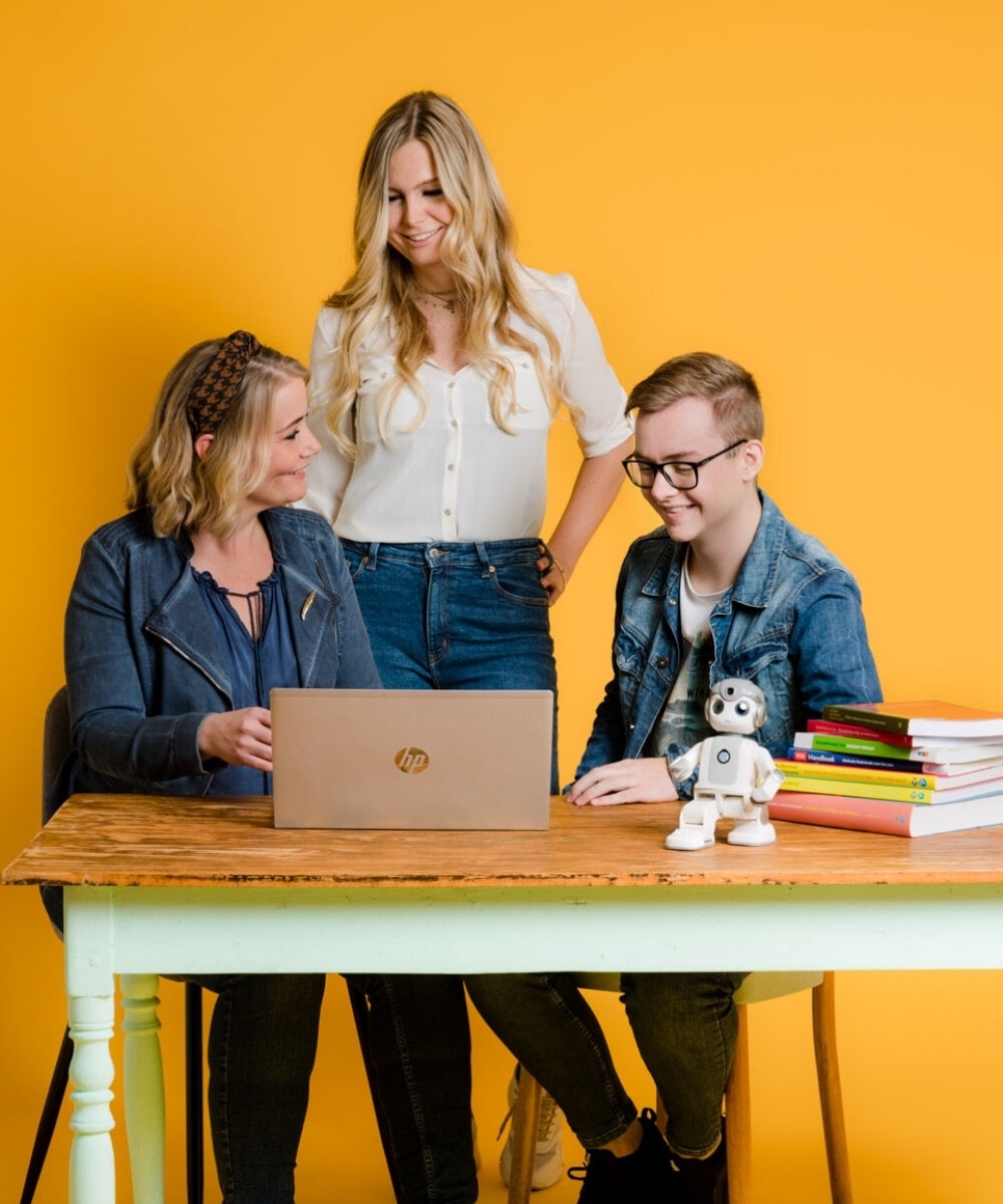 Twee lachende vrouwen en een lachende jonge man werken samen aan een laptop aan een tafel, met een kleine robot en een stapel boeken ernaast.