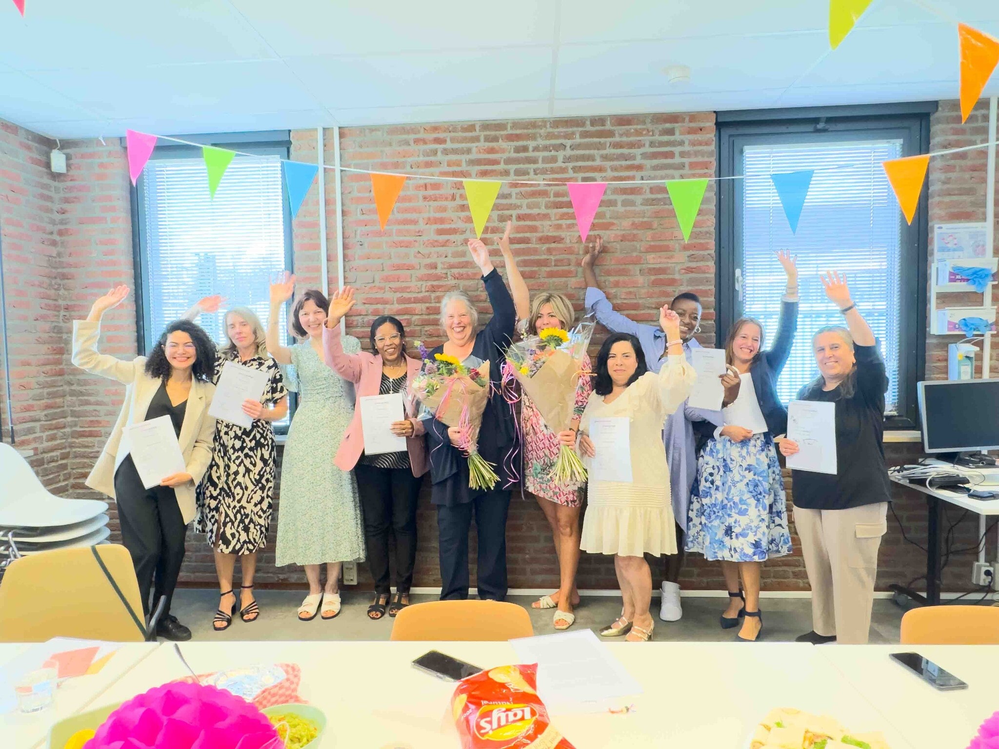 Tien feestelijk geklede vrouwen staan voor een bakstenen muur met kleurrijke slingers, houden certificaten vast en zwaaien, twee vrouwen hebben boeketten bloemen.