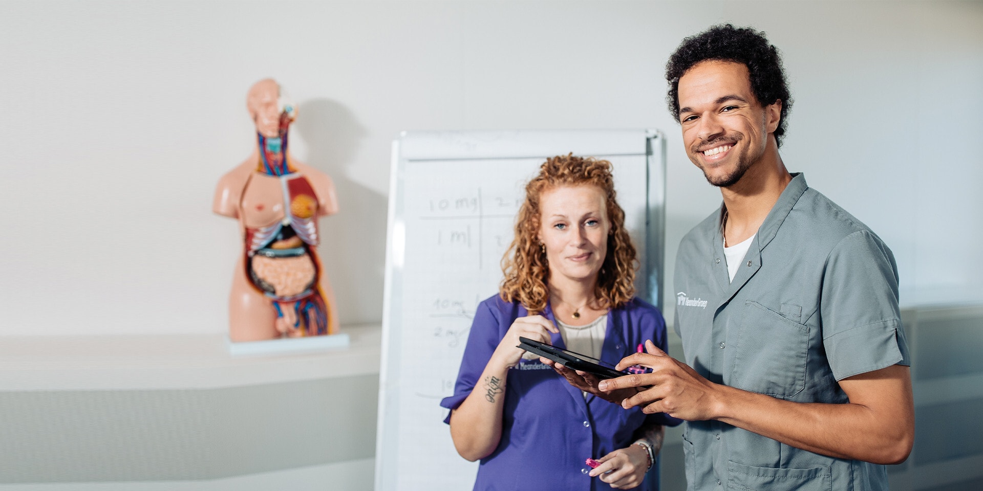 Twee glimlachende zorgmedewerkers, een man en een vrouw, staan in een klaslokaal met een tablet in hun handen.
