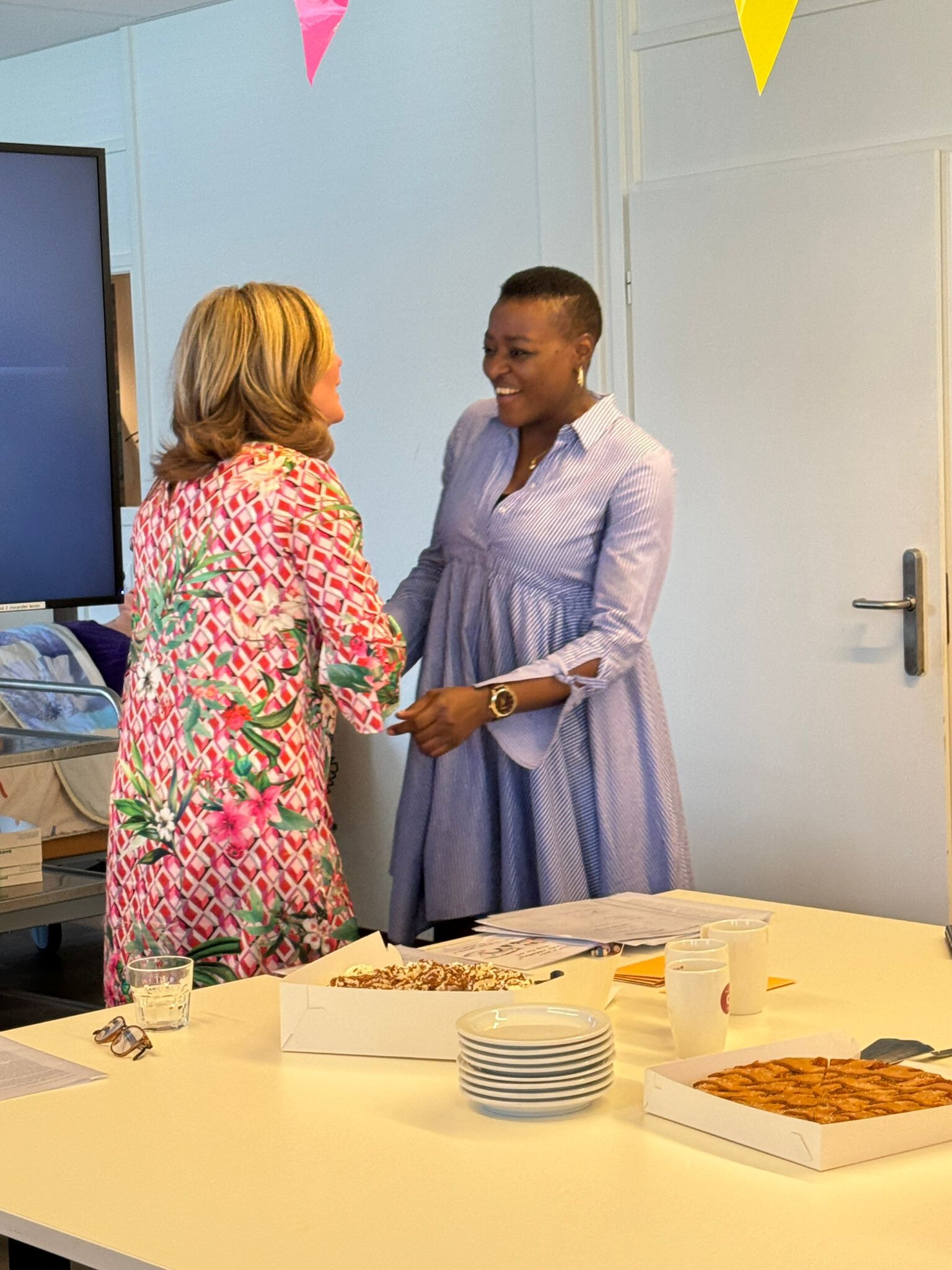 Twee vrouwen groeten elkaar lachend in een kamer met feestdecoraties, een tafel met gebak en servies op de voorgrond.