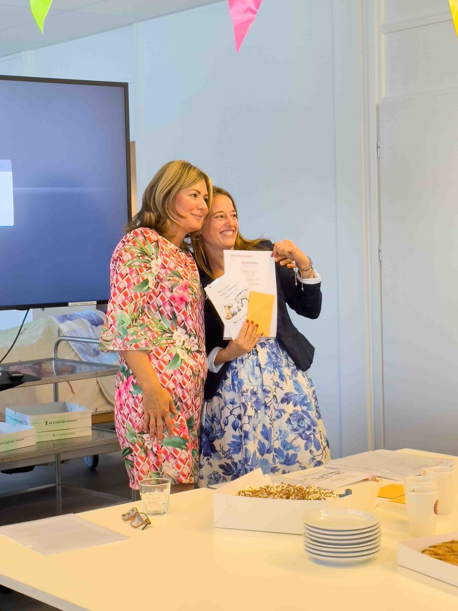 Twee vrouwen die samen poseren, één met een bloemenjurk en de ander met een blauwe bloemenrok en zwart jasje, waarbij de vrouw met de blauwe bloemenrok een certificaat en envelop vasthoudt, in een feestelijke setting met slingers en een tafel met taart en borden.