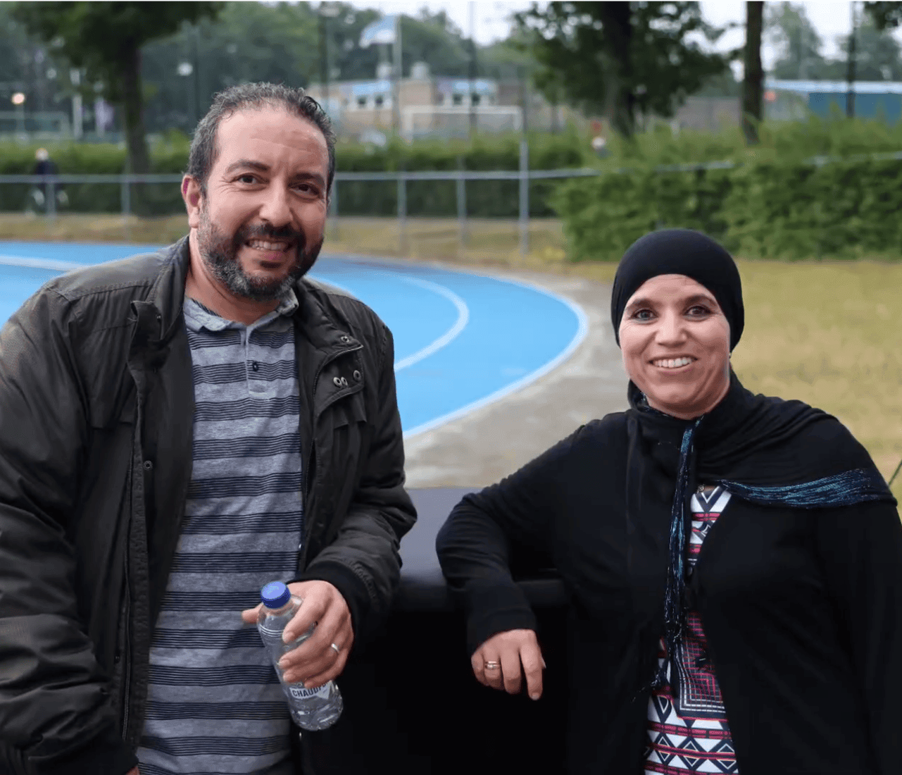 Lachende man en vrouw staan buiten op een sportveld, met de man die een flesje water vasthoudt.
