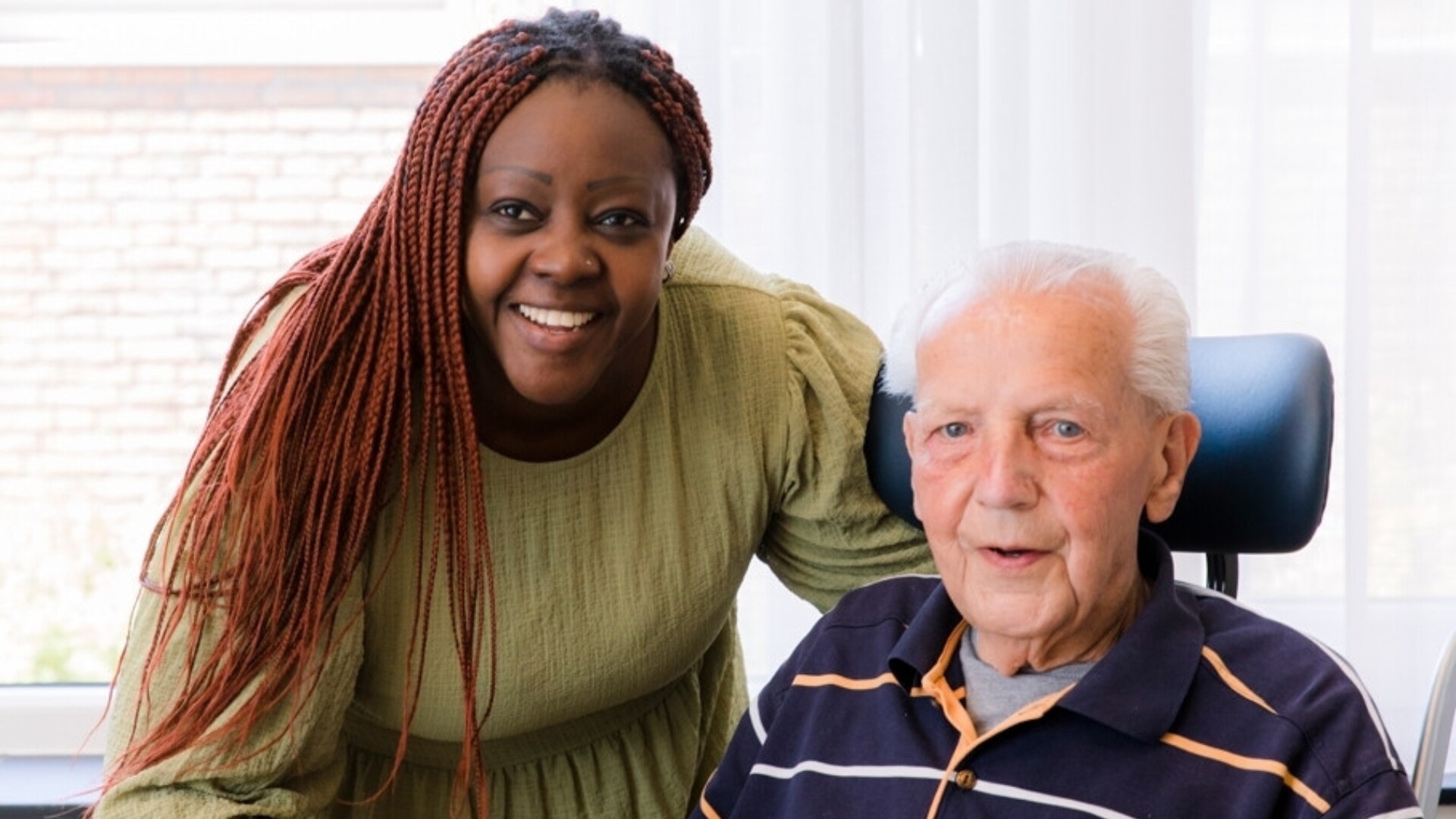 Een glimlachende, jonge vrouw staat achter een oudere man in een rolstoel.