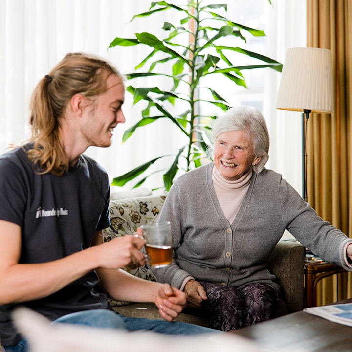 Een lachende oudere vrouw zit op de bank, terwijl een jonge man van MeanderGroep Thuis met een glas thee in zijn hand naar haar kijkt.