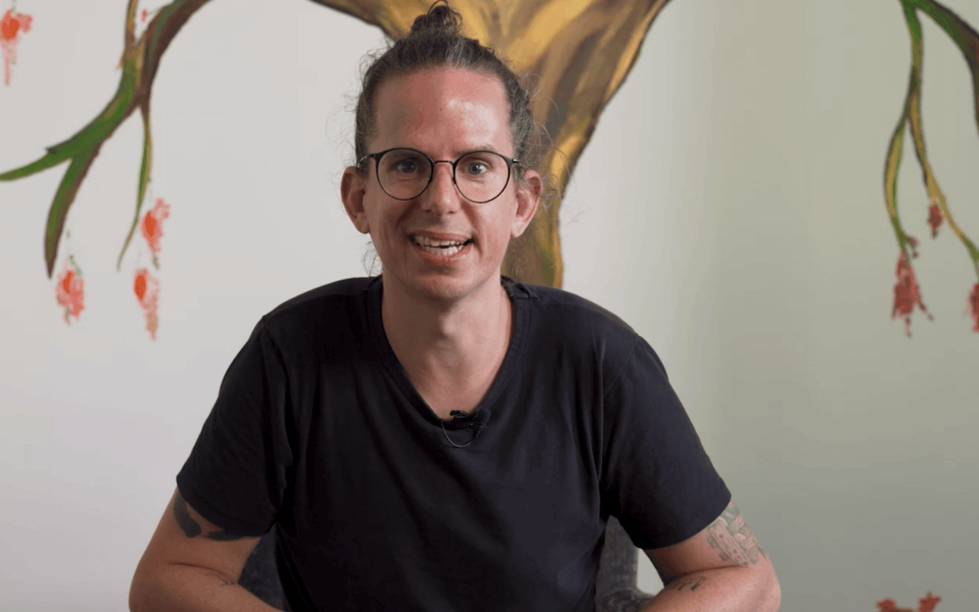 Persoon met lang haar in een knot, een bril en een zwart t-shirt, zittend voor een muur met een geschilderde boom en bloemen.
