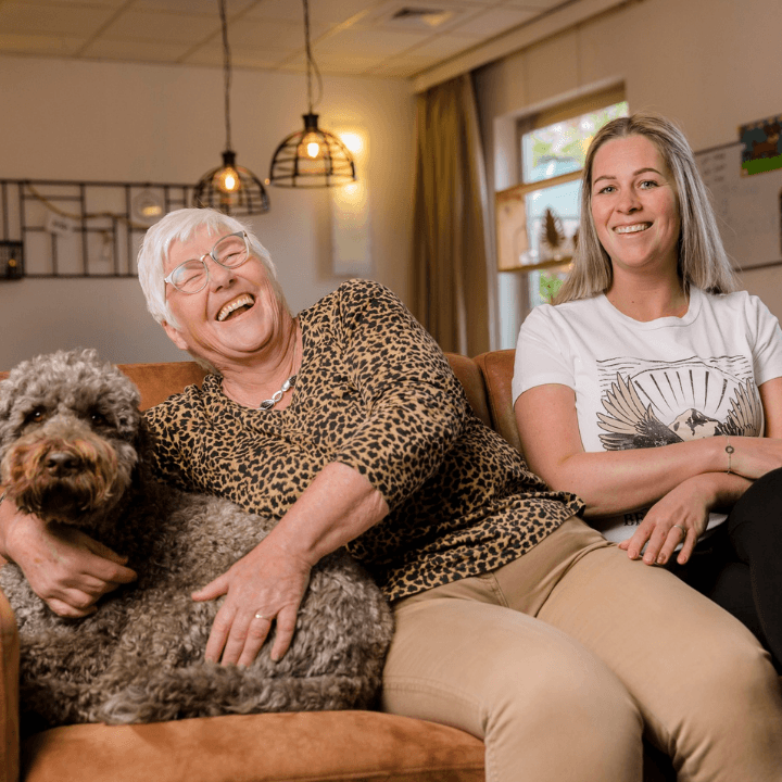 Lachende oude vrouw met hond zit op de bank naast een jonge vrouw.