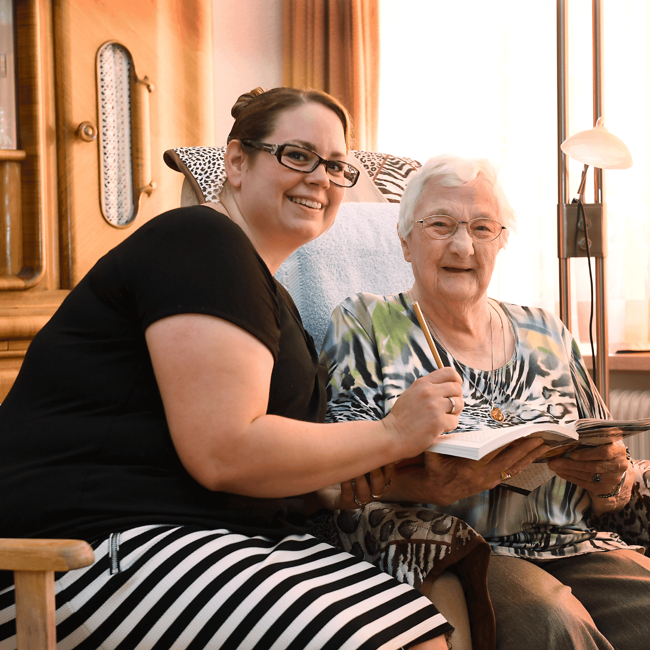Jonge vrouw met een zwart shirt en een gestreepte rok zit naast een lachende oude vrouw. Ze doen een kruiswoordpuzzel.