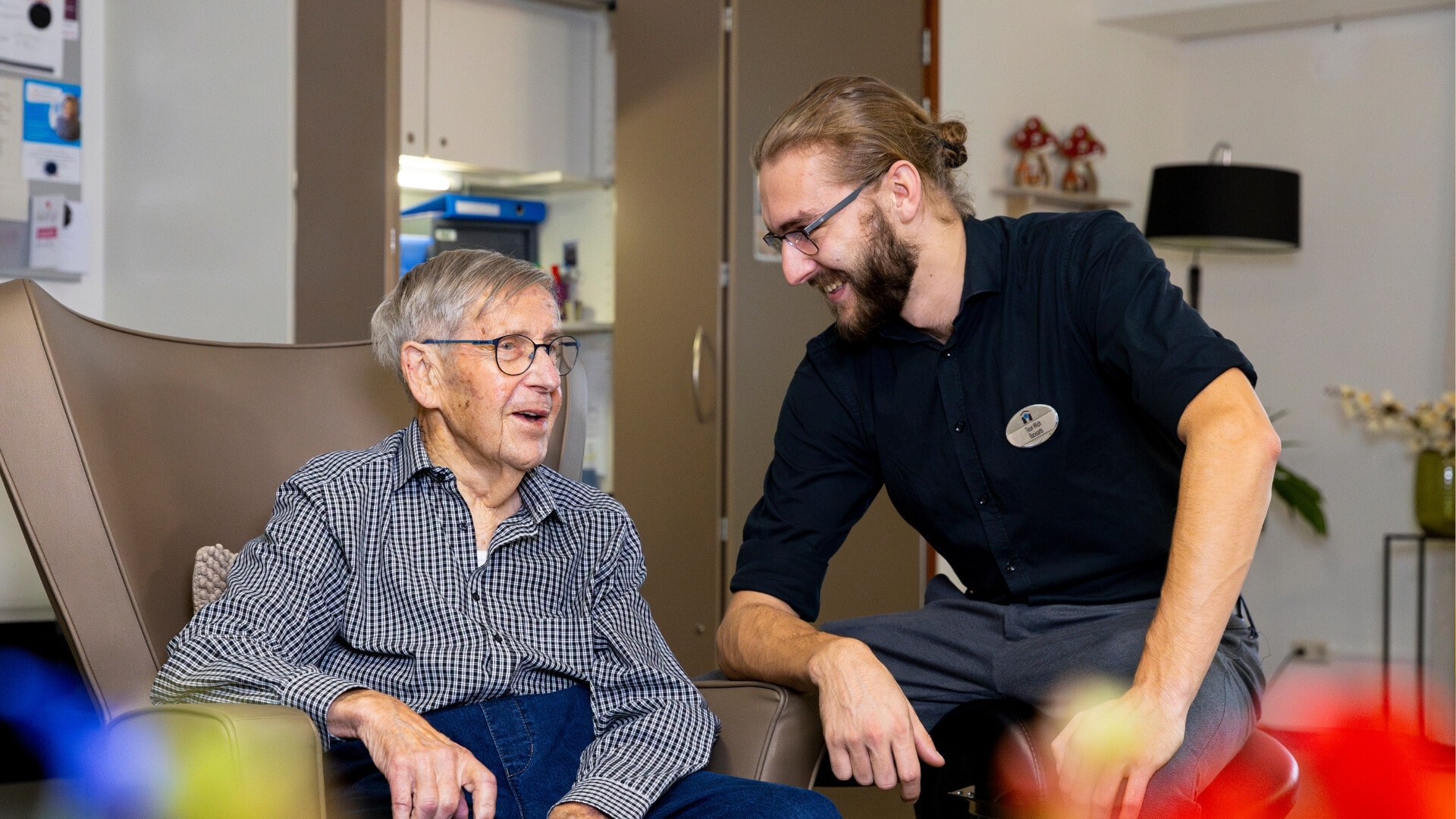 Oudere man met geruit hemd en bril zit op een stoel en praat met een jongere man die naast hem zit. De jongere man heeft een knotje, een bril en een zwart hemd aan. De jongere man lacht naar de oudere man.