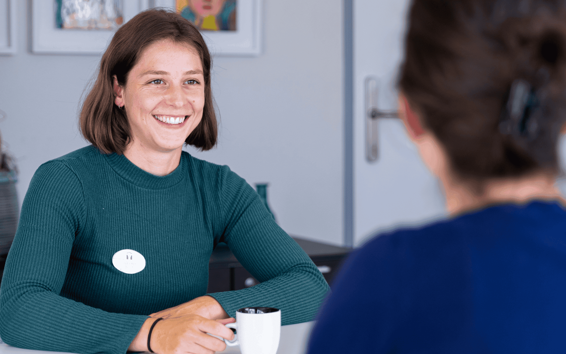 Een lachende vrouw met kort, bruin haar en een donkergroene trui zit aan een tafel. Ze heeft haar handen om een witte mok geklemd en is in gesprek met een vrouw wiens rug naar de camera is gekeerd.