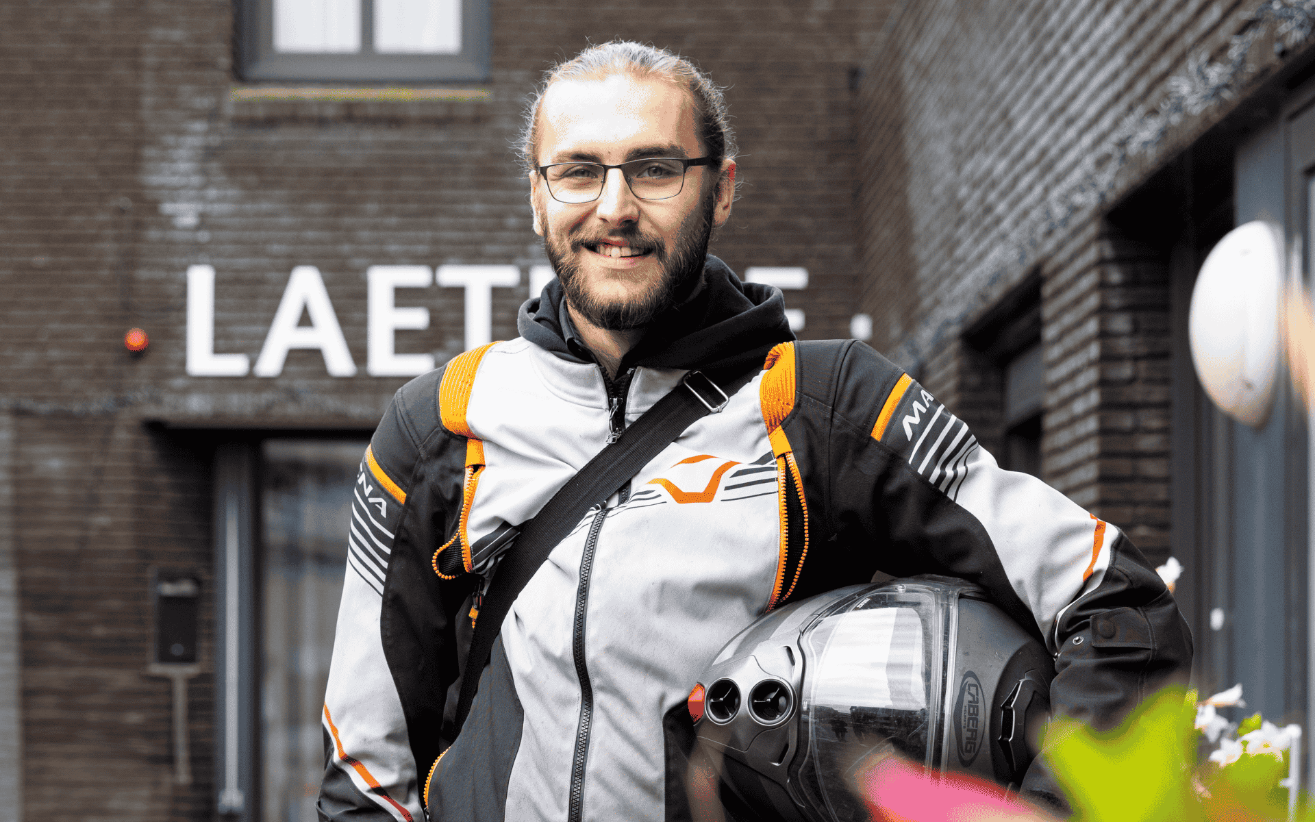 Lachende man met een baard en bril staat buiten. Hij draagt een grijze motorjas met oranje en zwarte accenten, een rugzak en houdt een motorhelm vast.