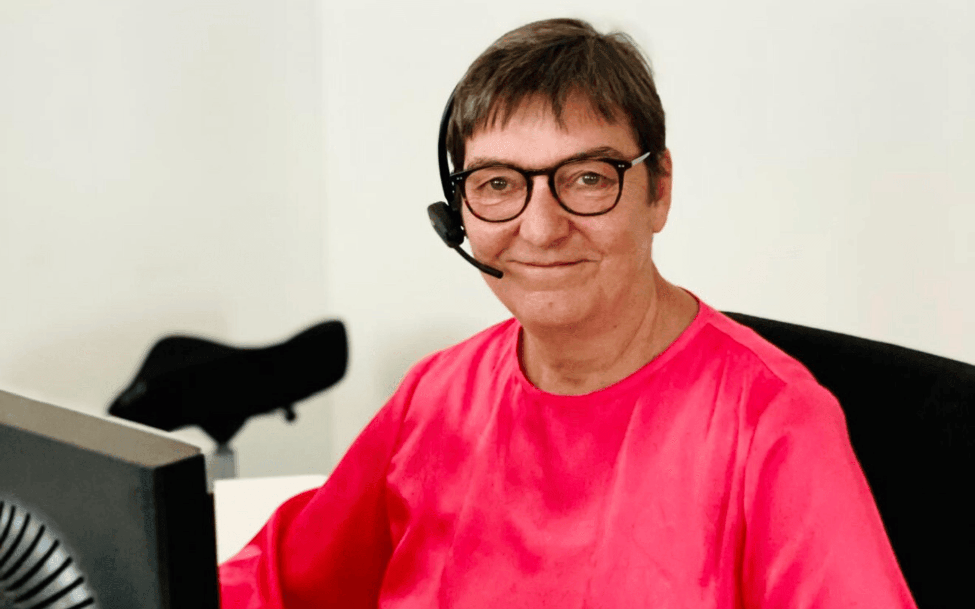 Portret van een glimlachende vrouw met kort haar, zwarte bril en een headset, zittend achter een computerscherm in een kantooromgeving.