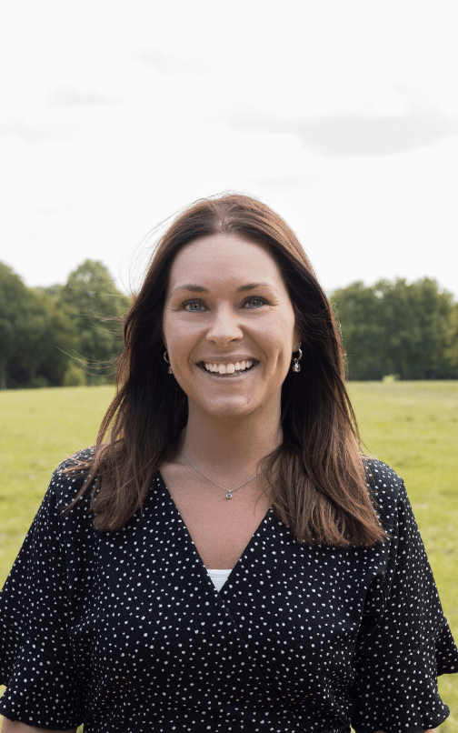 Lachende vrouw met lang donker haar en een zwart met wit gestipte blouse, staand voor een weiland.