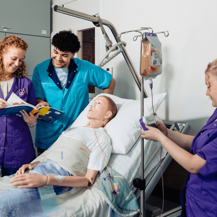 Zorgprofessionals in paarse en blauwe kleding verzorgen een medisch oefenpop in een ziekenhuisbed, een van hen bedient een infuus.