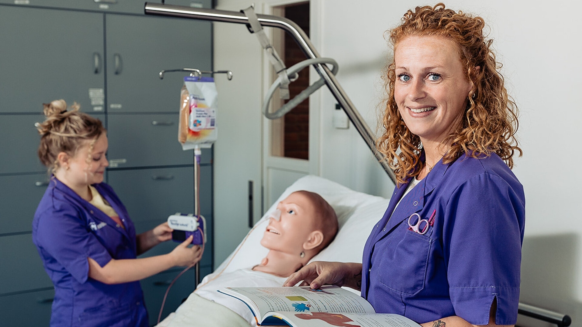 Twee vrouwelijke zorgmedewerkers staan naast een bed. In het bed ligt een medische oefenpop.