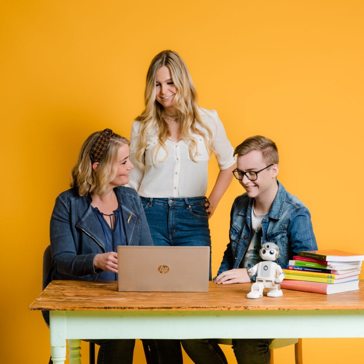Drie mensen zitten en staan rond een houten tafel met een laptop, een stapel boeken en een kleine witte robot, tegen een gele achtergrond.