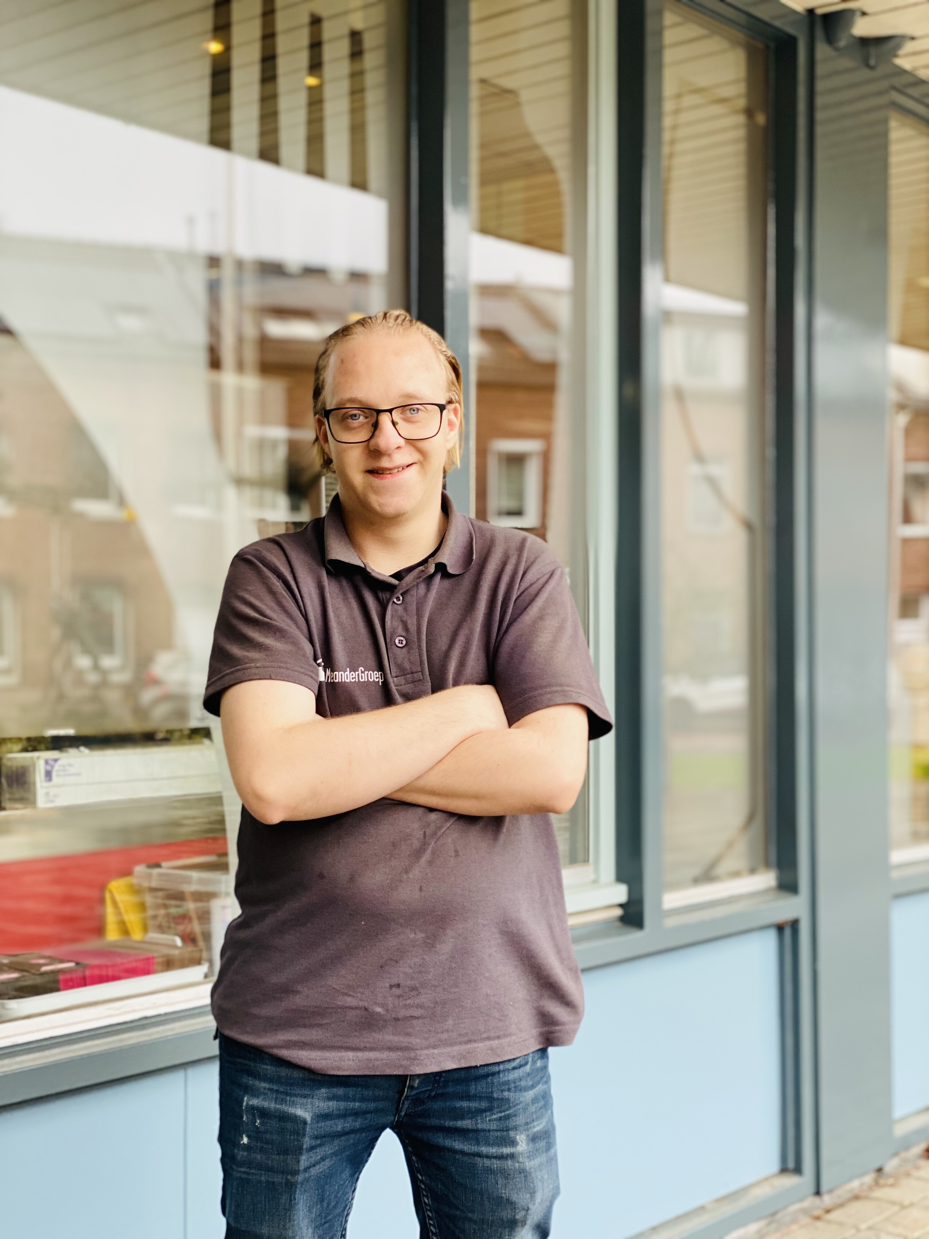 Een jonge man met een bril en een grijs poloshirt van MeanderGroep staat lachend buiten, met zijn armen over elkaar.