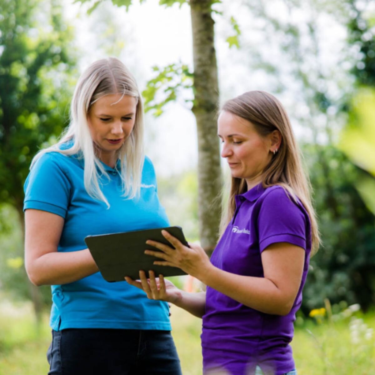Twee vrouwelijke medewerkers met een blauwe en een paarse polo kijken naar een ipad in het bos.