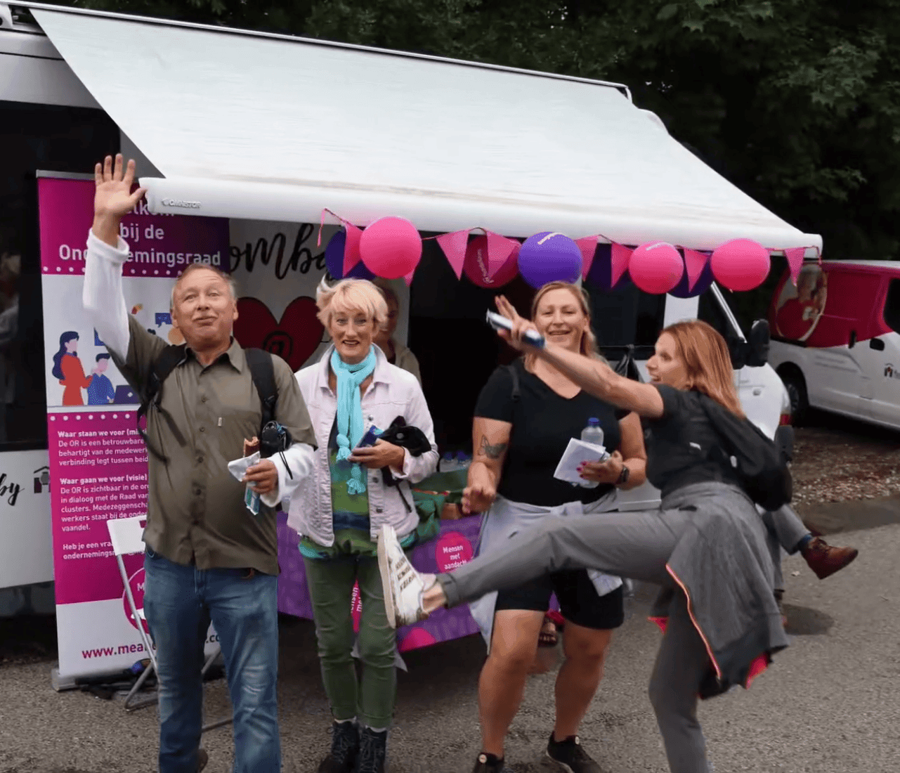 Vier lachende collega's staan buiten bij een kraam met roze en paarse lampionnen. Een vrouw maakt een speelse schopbeweging.