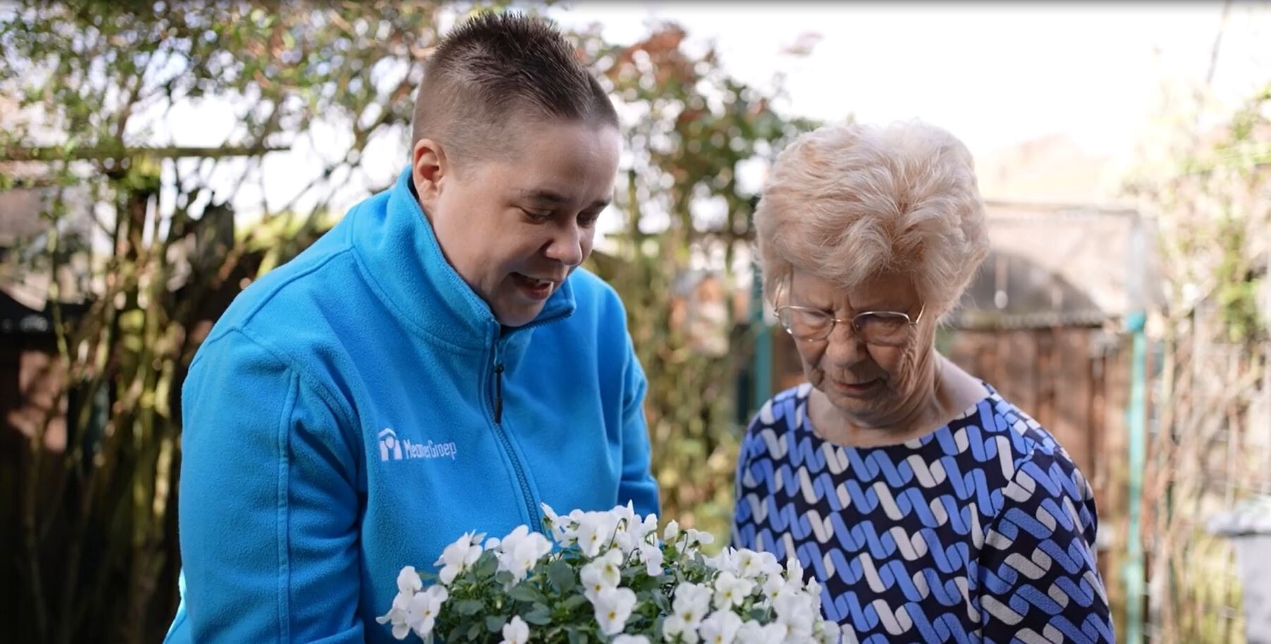 Medewerker van MeanderGroep staat in een tuin met een oudere vrouw. Ze houden een plant vast met witte bloemen.