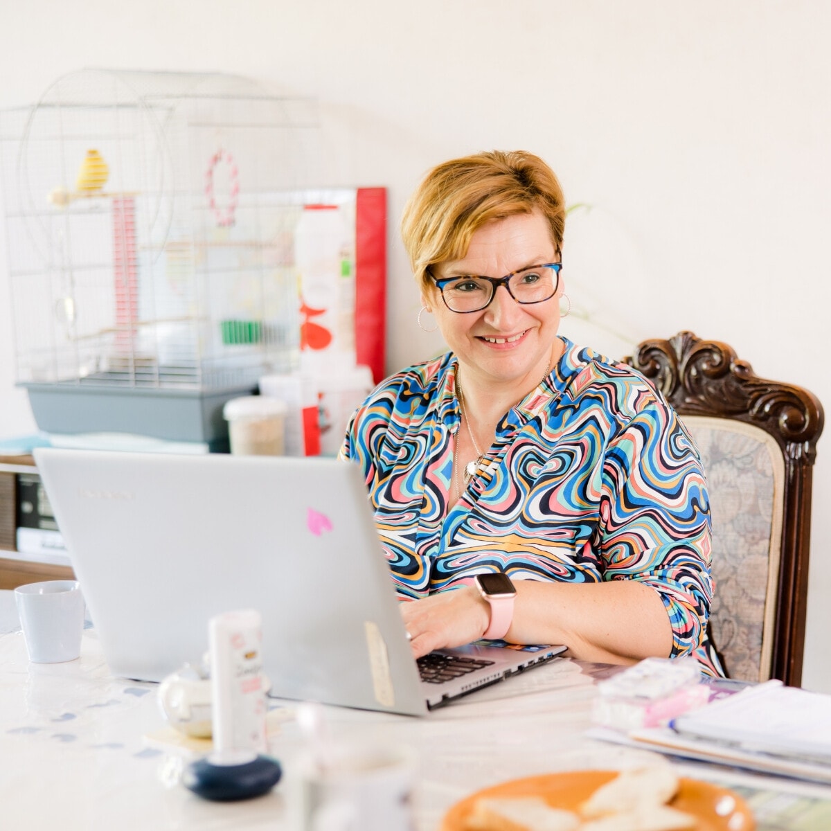 Een lachende vrouw met een bril en kleurrijke blouse zit aan een tafel en werkt op haar laptop. In de achtergrond staat een vogelkooi met een parkiet.